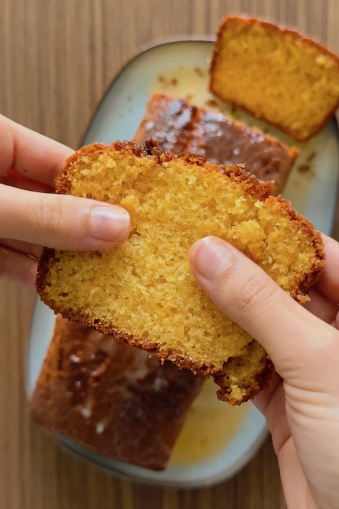 Close-up of hands holding a soft, moist slice of gluten-free orange cake showing the light, porous crumb and orange zest.